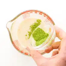A hand pours matcha powder into a measuring cup with smoothie mixture.