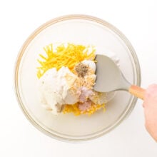 A hand holds a spatula, stirring together vegan cheese and other ingredients in a bowl.