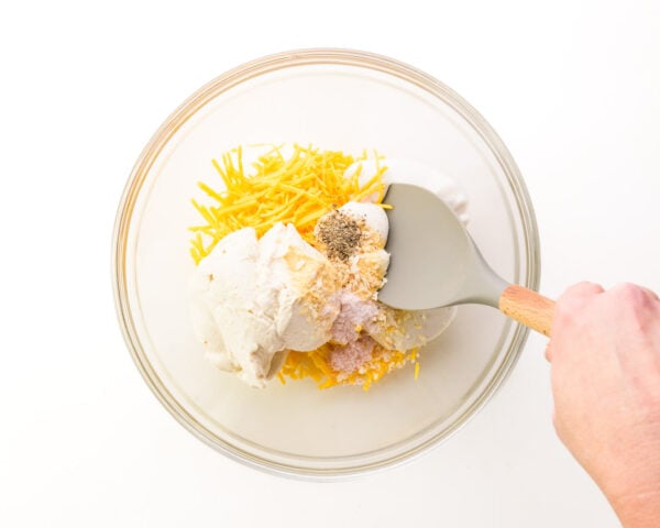 A hand holds a spatula, stirring together vegan cheese and other ingredients in a bowl.
