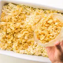 A buttery cornflakes mixture is being poured over potatoes in a baking dish.