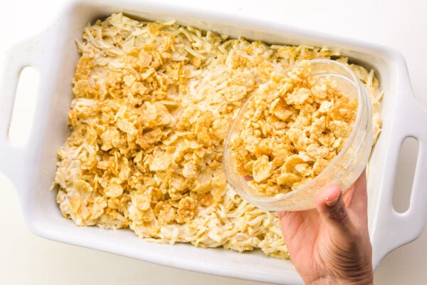 A buttery cornflakes mixture is being poured over potatoes in a baking dish.
