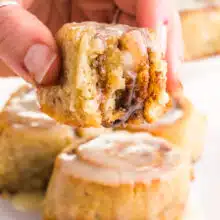 A hand holds a cinnamon roll with a bite taken out, hovering over more cinnamon rolls.