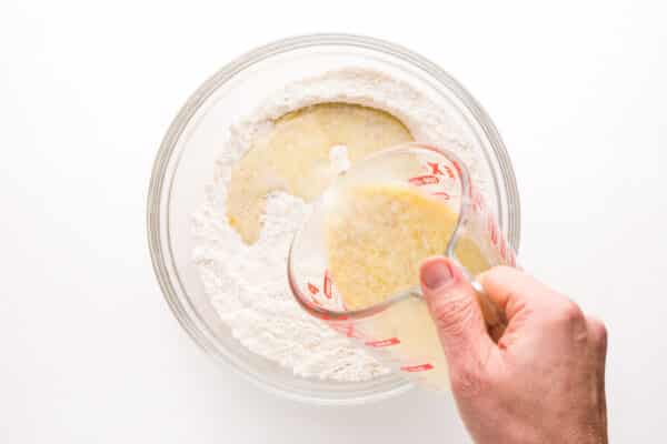 A hand holds a pyrex measuring dish, pouring yeast liquid into a flour mixture.
