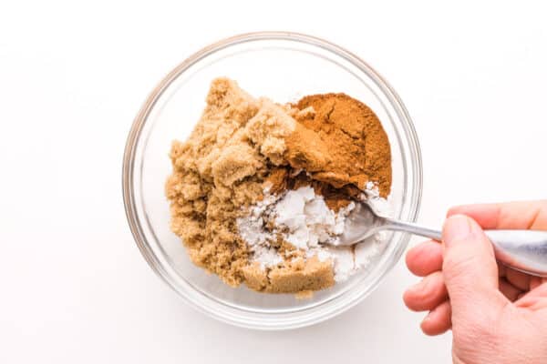 A hand holds a spoon, stirring cinnamon, brown sugar, and other ingredients in a bowl.