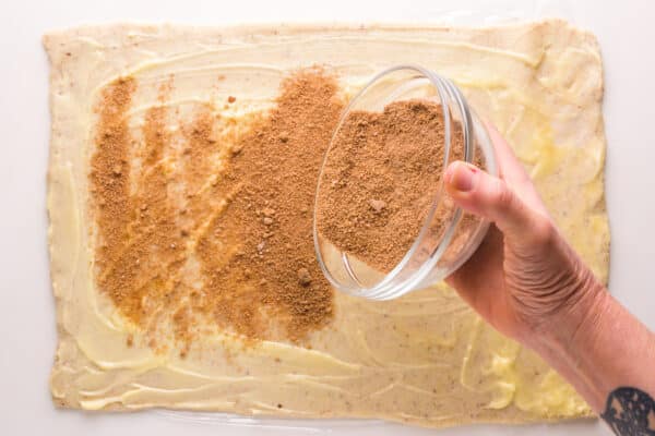 A hand holds a bowl with a cinnamon mixture, sprinkling it over rolled out dough.