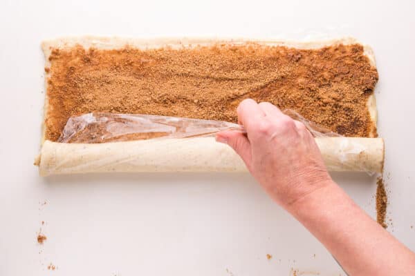 A hand holds plastic wrap, using it to roll a long sheet of dough sprinkled with cinnamon sugar.