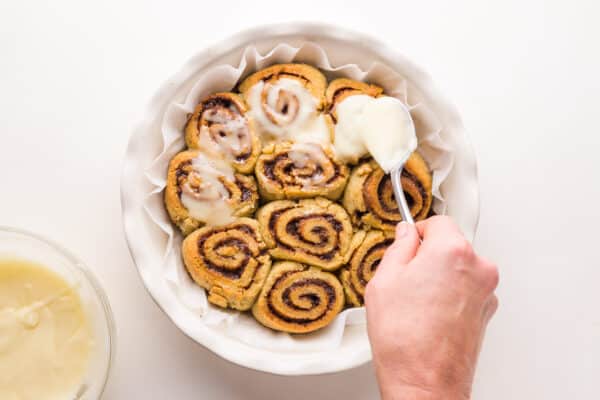 A hand holds a spoon, adding icing to freshly baked cinnamon rolls.