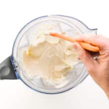 A hand holds a spatula, scraping down the sides of a blender jar with a creamy white mixture in it.