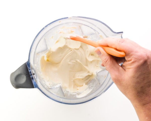 A hand holds a spatula, scraping down the sides of a blender jar with a creamy white mixture in it.