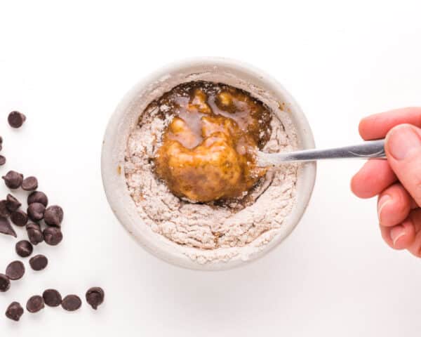 A hand holds a spoon, stirring ingredients in a bowl. There are chocolate chips on the side.