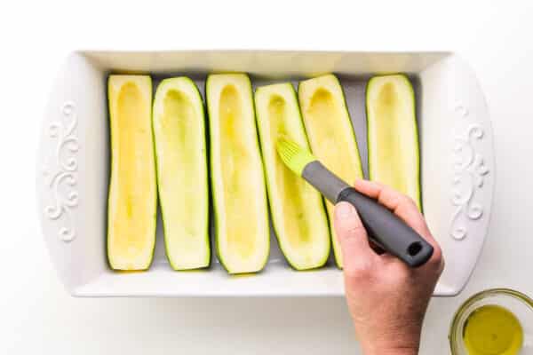 A hand holds a melon scooper, scooping flesh out of zucchinis in a baking dish.