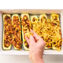 A hand sprinkles vegan cheese over zucchini boats in a baking dish.