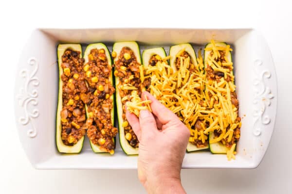 A hand sprinkles vegan cheese over zucchini boats in a baking dish.