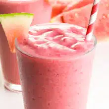 A watermelon smoothie consists of a paper straw and wedges of watermelon.  In the background is another smoothie and a slice of watermelon.