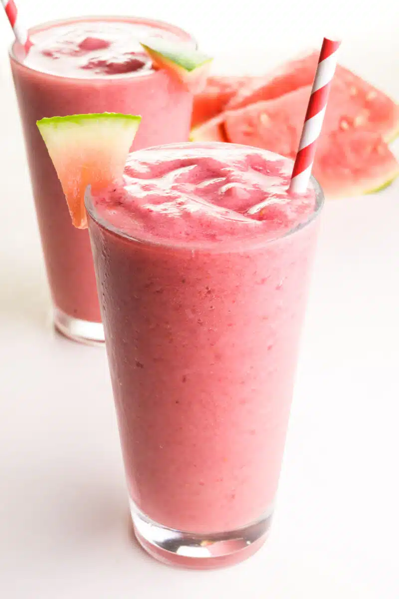 Two pink smoothies are sitting on a table.  They have watermelon wedges and paper straws.  There are watermelon wedges in the background.