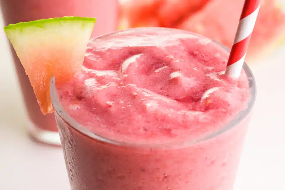 A closeup of a watermelon banana smoothie in a glass  It has a red and white straw and watermelon wedge.