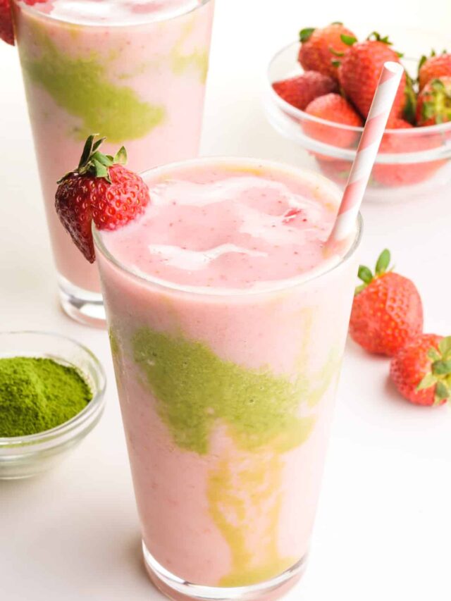Pink and green smoothie liquid is in two glasses, one in front of the other. There is a bowl of green tea powder and strawberries around the glasses.