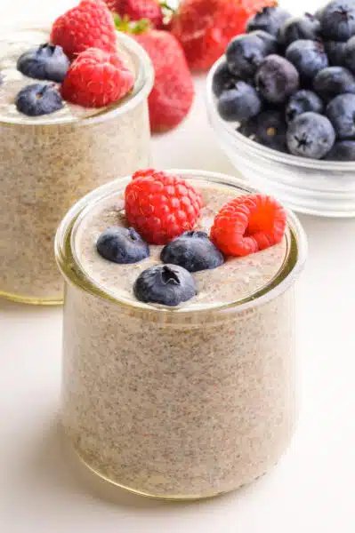A glass dish contains chia flax pudding with blueberries and raspberries.  It sits in front of another bowl of pudding and fresh berries.