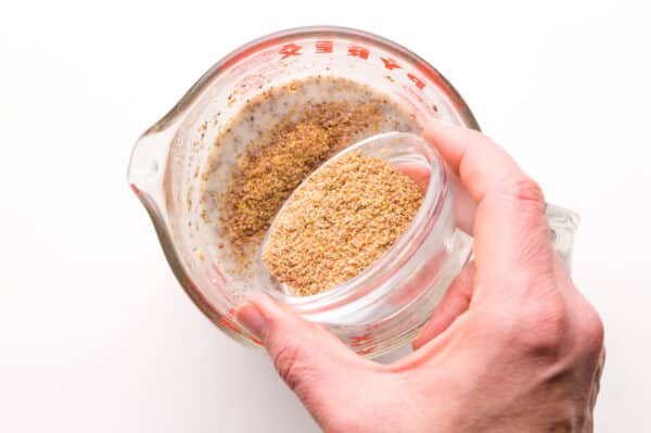 A hand holds a bowl of ground flax, pouring into a bowl with chia pudding.