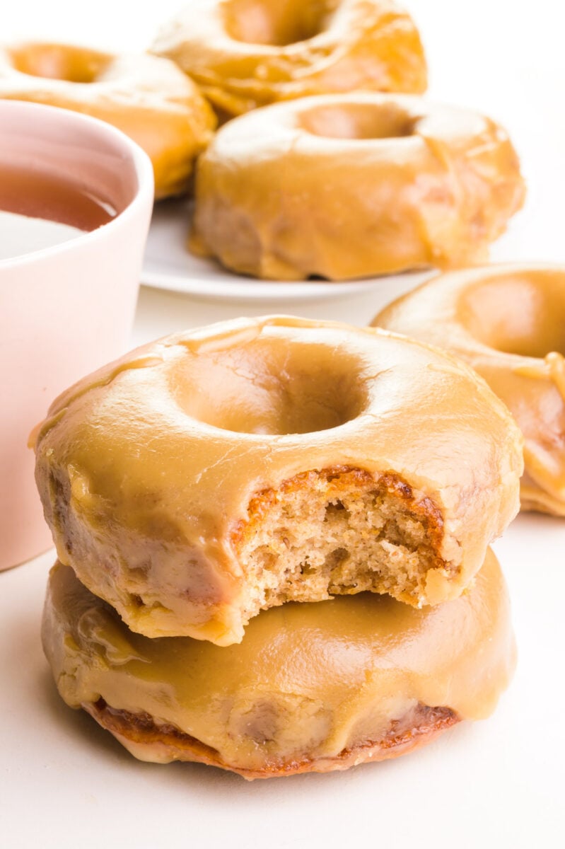 A stack of maple donuts shows the top one with a bite taken out. The stack sits next to a pink mug of tea. There are more donuts in the background.