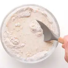 A hand holds a spatula, stirring flour with milk ingredients in a glass mixing bowl.