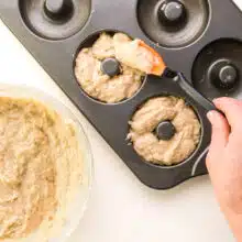 A hand uses a spatula to spread donut batter into a donut pan.