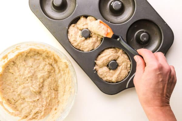 A hand uses a spatula to spread donut batter into a donut pan.