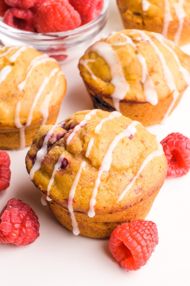 Several muffins sit next to fresh raspberries. The muffins have drizzles of icing on top.
