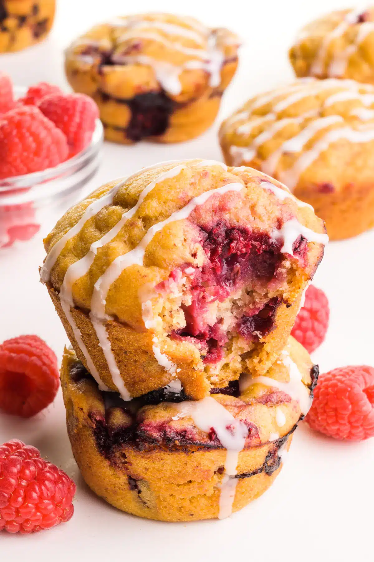 A stack of muffins shows the top one with a bite taken out. There are fresh raspberries and more muffins around it.