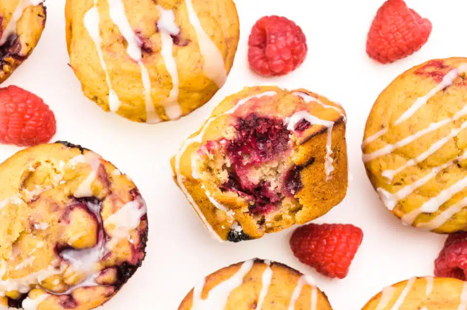 Looking down on several muffins with fresh raspberries nearby. There is one muffin with a bite taken out, showing lots of raspberries inside.