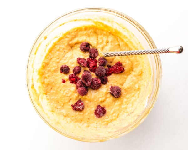 Looking down on a bowl of muffin batter with raspberries in the center. There is a spoon for stirring in the batter too.