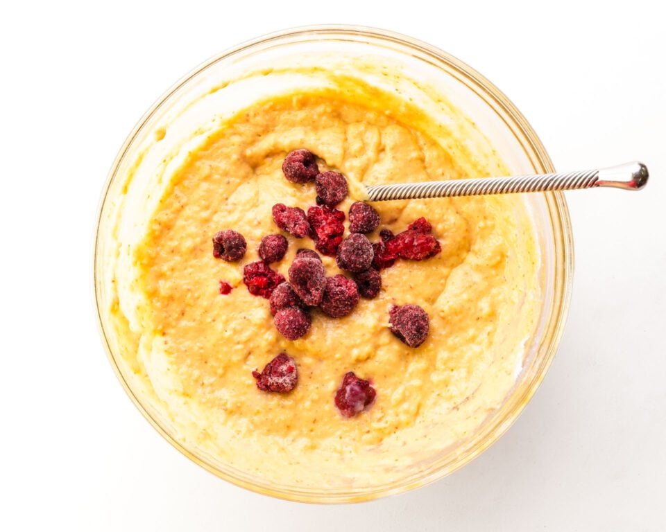 Looking down on a bowl of muffin batter with raspberries in the center. There is a spoon for stirring in the batter too.