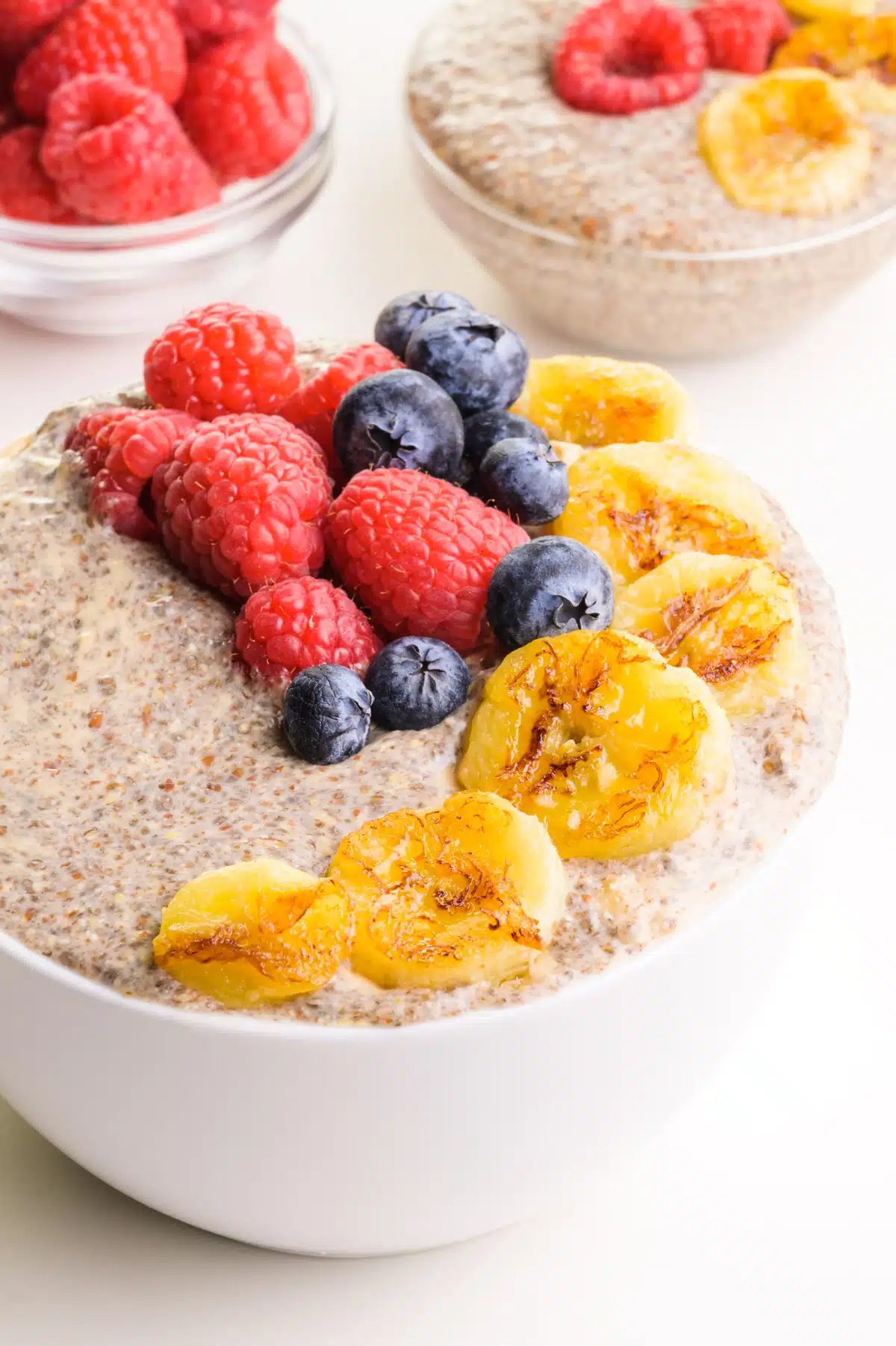 A bowl of warm chia pudding has seared bananas and fresh berries on top. There is another bowl of the pudding and a bowl of fresh raspberries in the background.