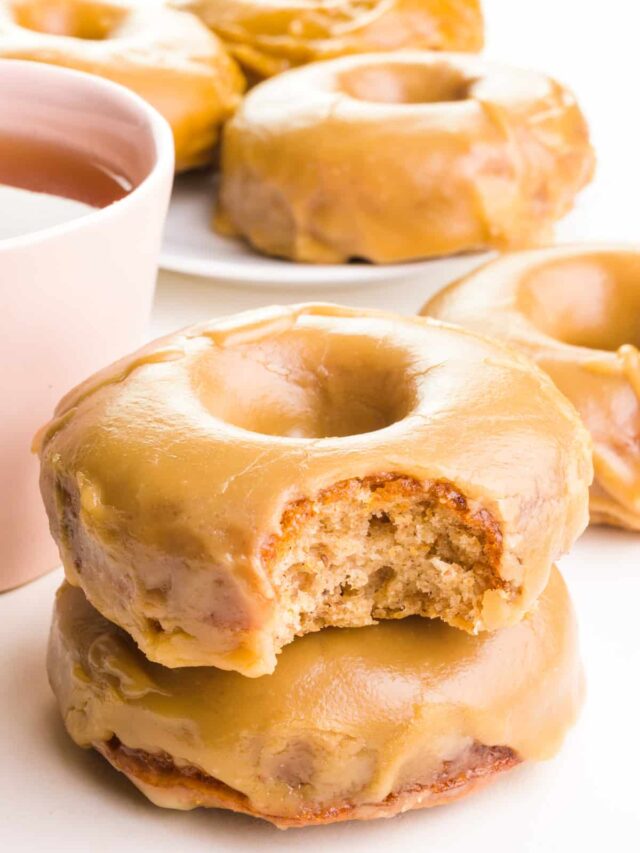 A stack of maple donuts shows the top one with a bite taken out. The stack sits next to a pink mug of tea. There are more donuts in the background.