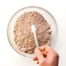 A hand holds a measuring spoon of baking powder over a bowl with boxed brownie mix in it.