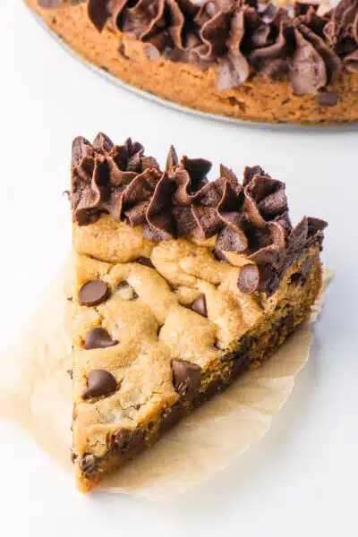 A slice of chocolate chip cookie cake on a napkin sits in front of the rest of the cake.