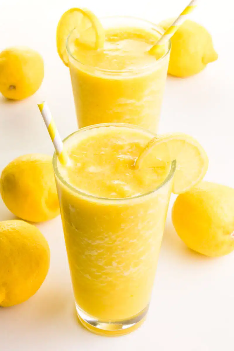 Two glasses of smoothies hold bright orange mixtures. There are lemon wedges and straws in the glasses. There are fresh lemons around the smoothies.