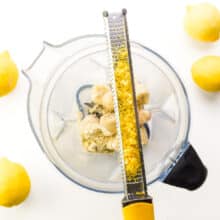 Lemon zest sits in a microplane on top of a blender with other ingredients. There are fresh lemons around the blender.