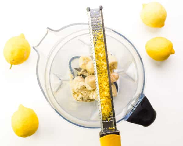 Lemon zest sits in a microplane on top of a blender with other ingredients. There are fresh lemons around the blender.