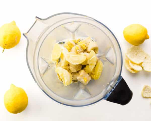 Looking down on a blender with fresh lemon pieces and other ingredients. There are fresh lemons and lemon peels around the blender.
