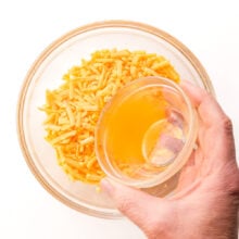A hand holds a bowl of vegetable broth, pouring it into a bowl with vegan cheddar shreds.