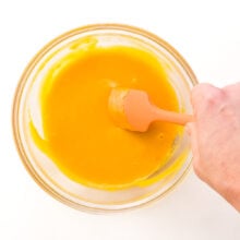 A hand holds a rubber spatula, stirring melted vegan cheese in a bowl.