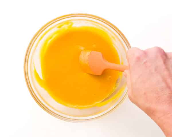 A hand holds a rubber spatula, stirring melted vegan cheese in a bowl.