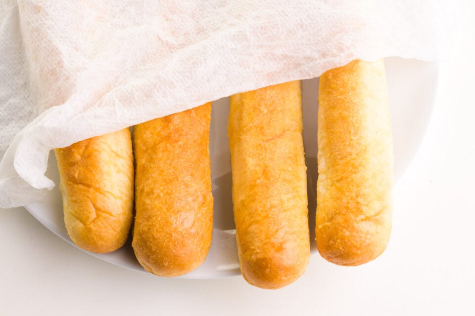 Breadsticks are on a plate and topped with a moistened paper towel, waiting to be reheated in a microwave.