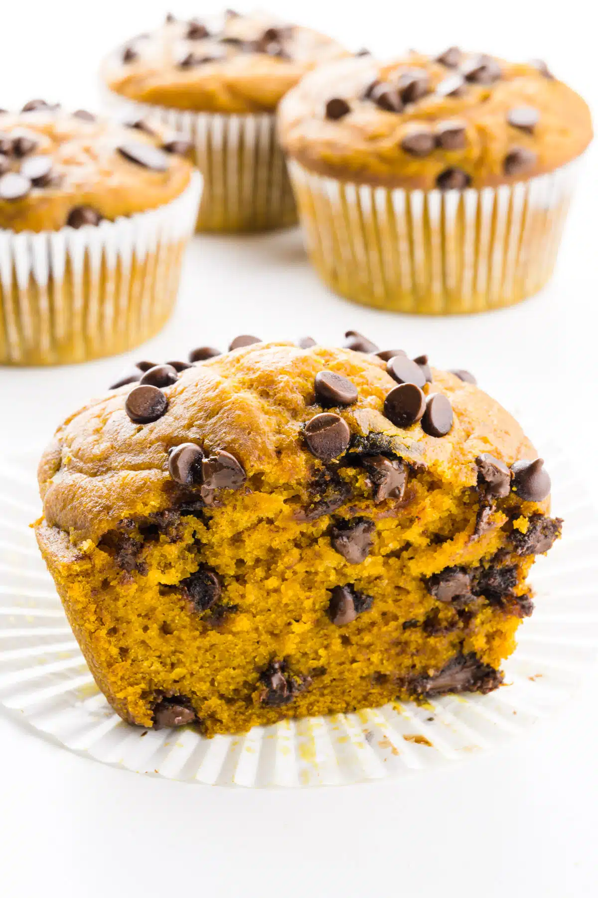 A muffin is cut in half showing chocolate chips in the middle and on top. There are more muffins behind it.