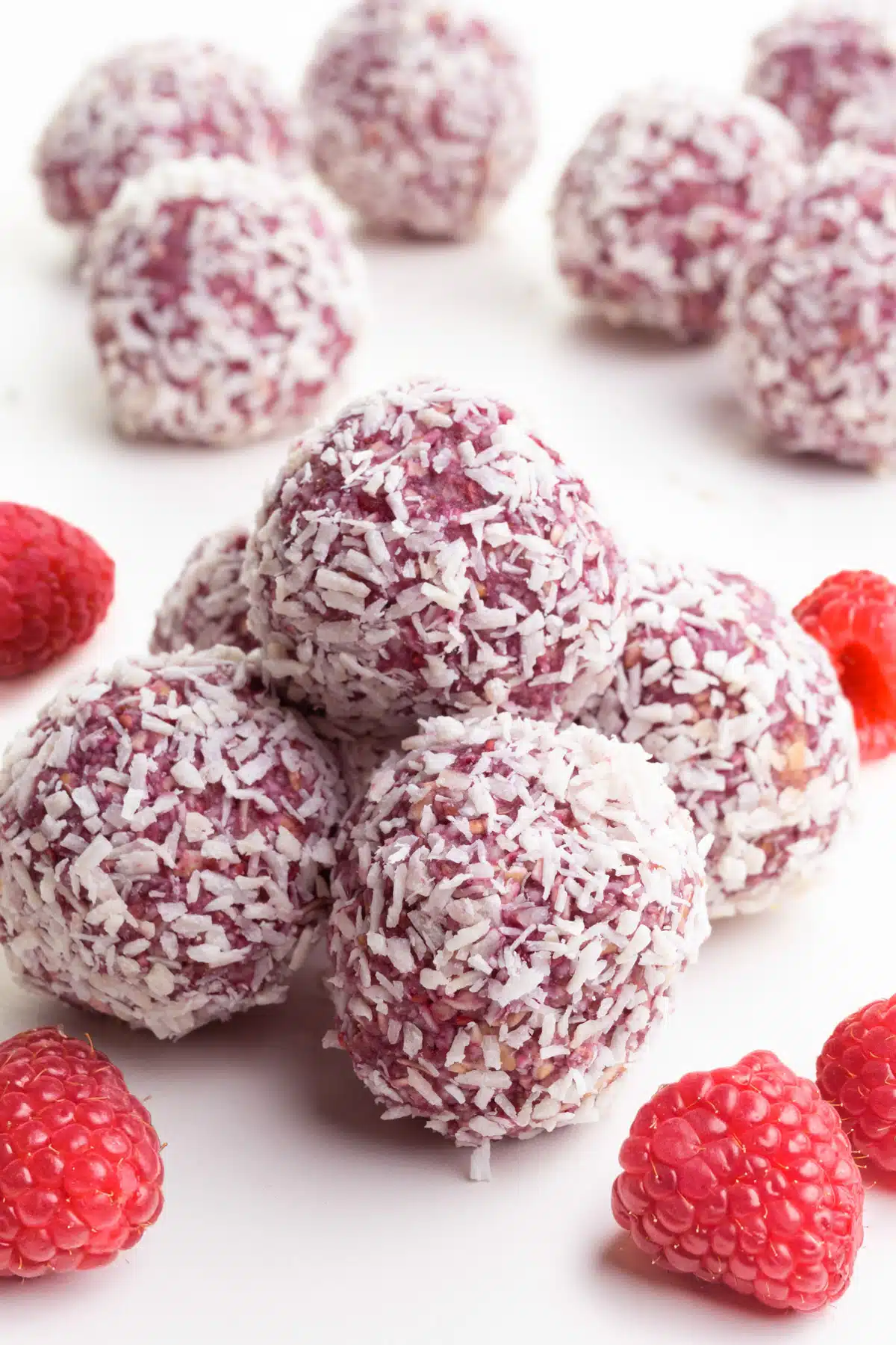 A stack of raspberry coconut bliss balls sits near fresh raspberries and more of the treats in the background.