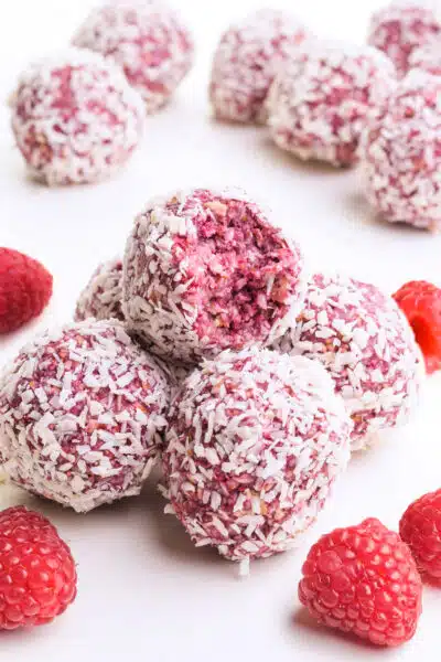 A stack of raspberry bliss balls shows the top one with a bite taken out. There are raspberries around the stack and more bliss balls in the background.