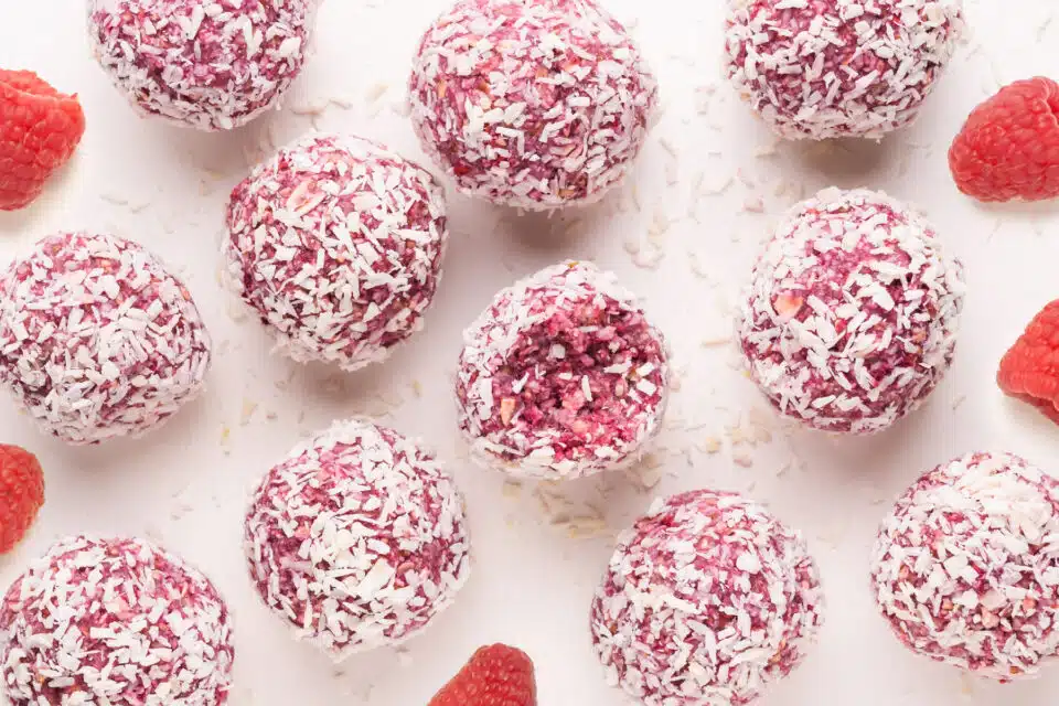 Looking down on raspberry coconut balls on a white table. The middle one has a bite taken out. There are fresh raspberries scattered around the treats.