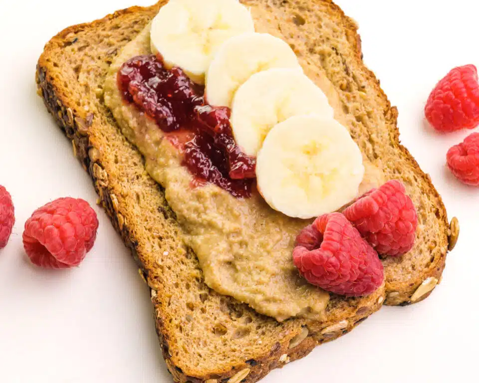 Looking down on a slice of bread with tigernut butter, fresh fruit, and jelly. There are fresh raspberries around it.