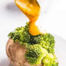 A spoon is drizzling vegan cheese sauce over a baked potato with steamed broccoli. A bowl of more cheese sauce is sitting in the background.
